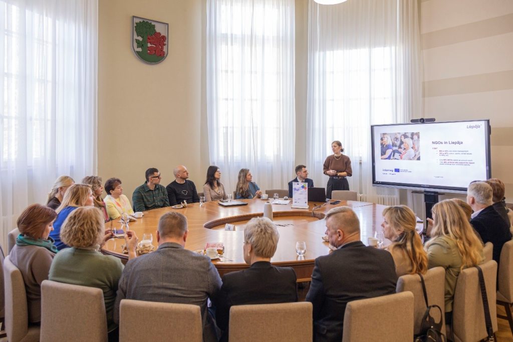 Meeting in a conference room with participants engaged in a presentation about NGOs in Liepāja, featuring a speaker addressing the audience and a large screen displaying information. The setting includes a round table, chairs, and a coat of arms on the wall.