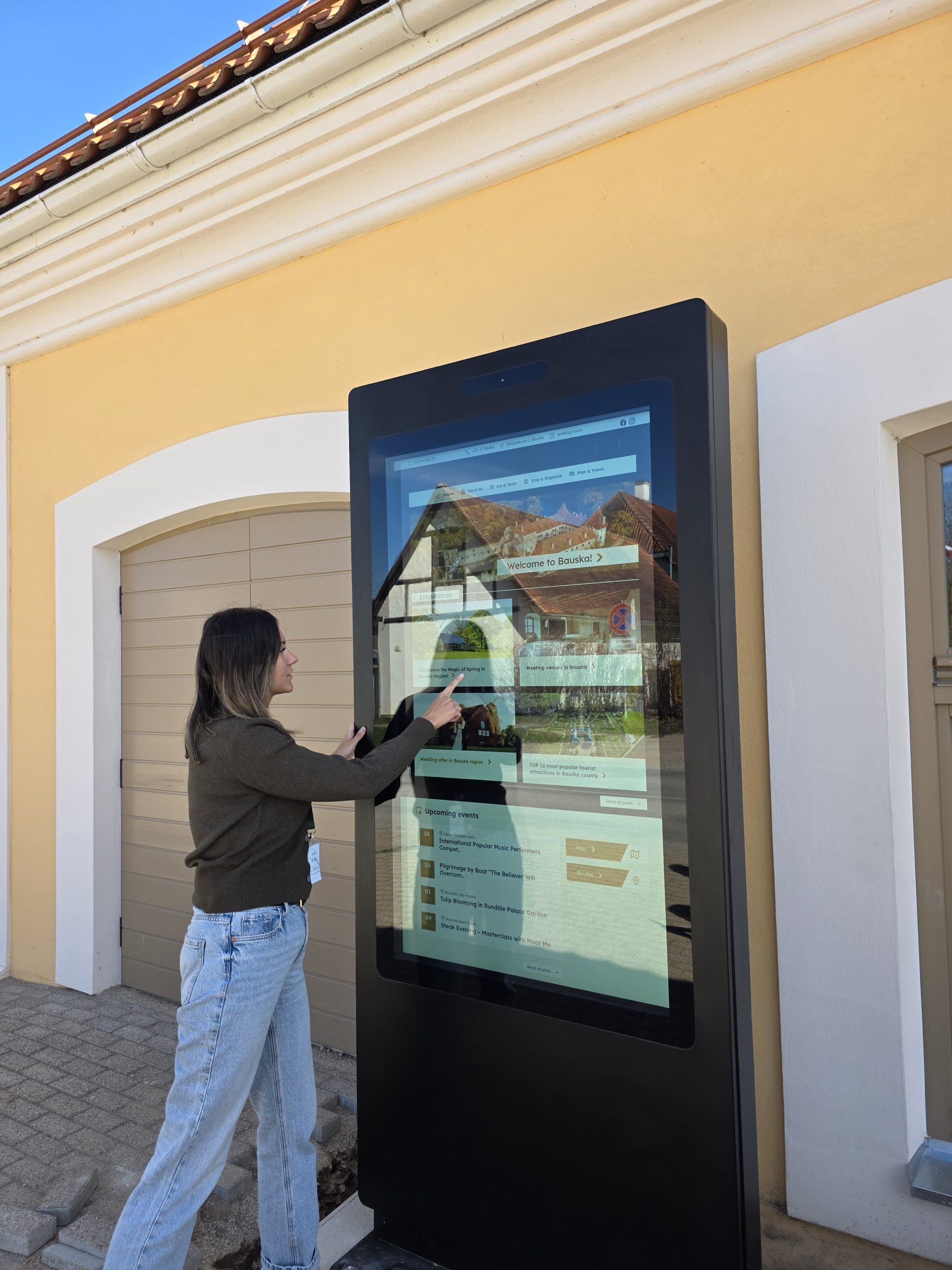 A woman interacts with a digital information kiosk displaying events and attractions in Bauska, set against a yellow building. The kiosk features a user-friendly interface for visitors.
