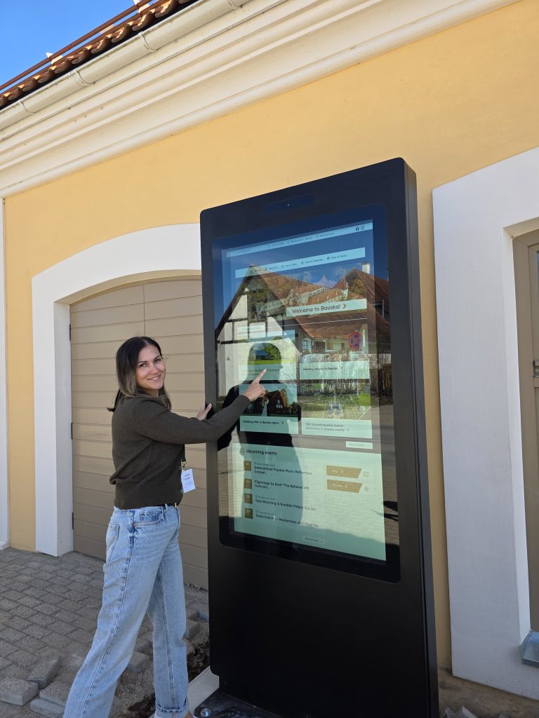 Young woman interacting with a digital information kiosk in Bauska, Latvia, displaying local events and attractions against a bright blue sky and yellow building backdrop.