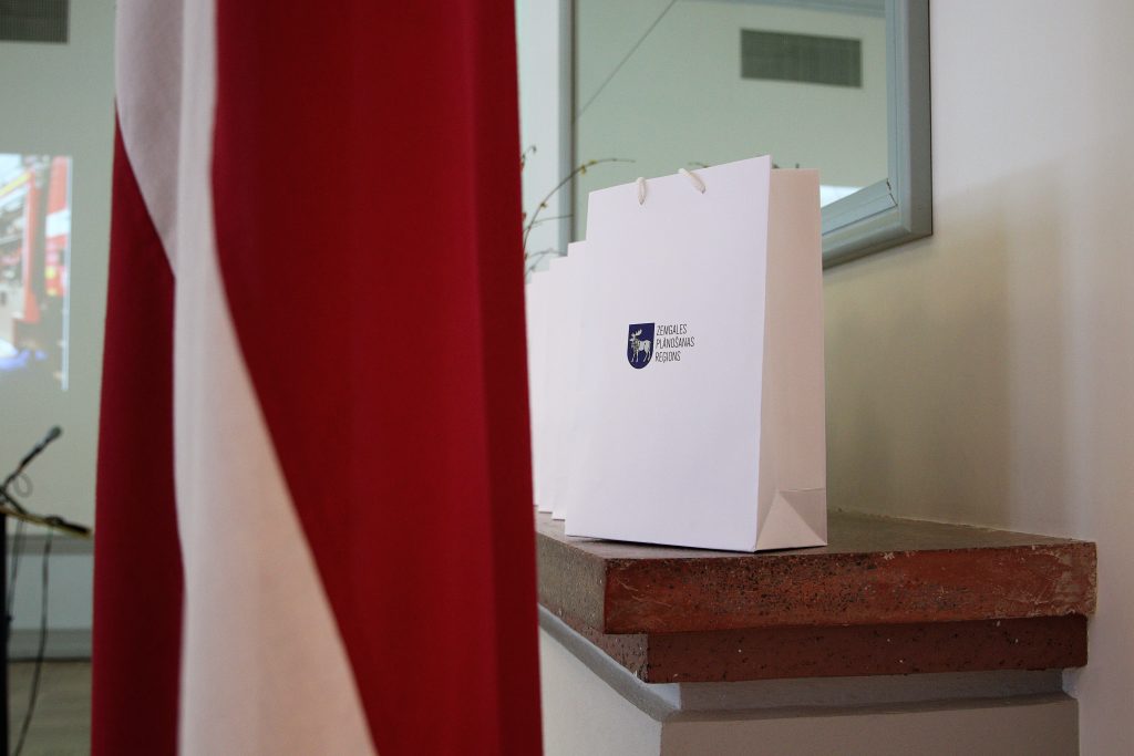 White gift bags with a logo on a stone ledge, partially obscured by a red flag, in a conference setting.