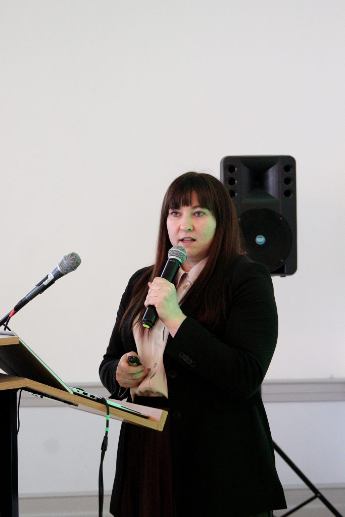 A female speaker presenting at a conference, with a sound system visible in the background.