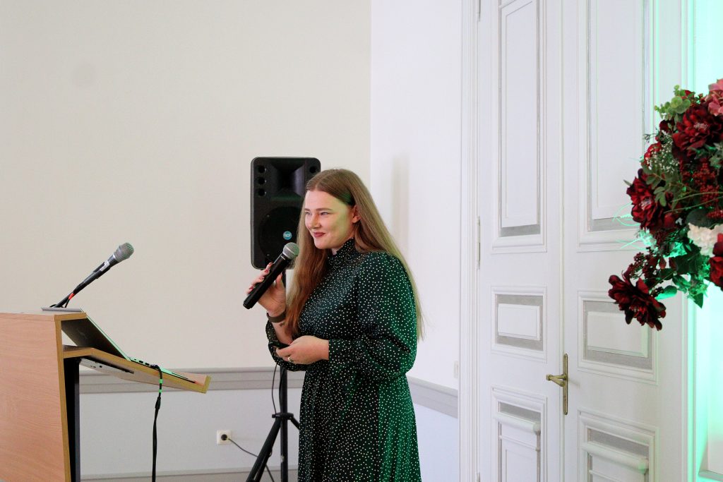 Young woman speaking at a podium with a microphone in a formal setting, dressed in a polka-dot dress, with a speaker and floral arrangement in the background.