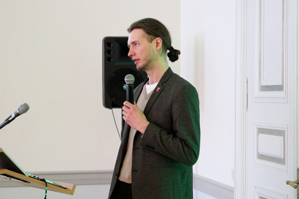 Man speaking at a podium with a microphone in a conference setting, wearing a blazer and holding a microphone, with a speaker visible in the background.