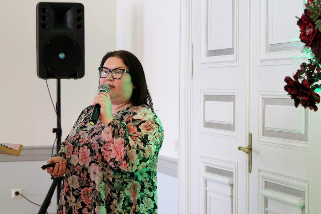 Woman with glasses speaking into a microphone at an event, wearing a floral-patterned blouse, with a speaker and a door in the background.
