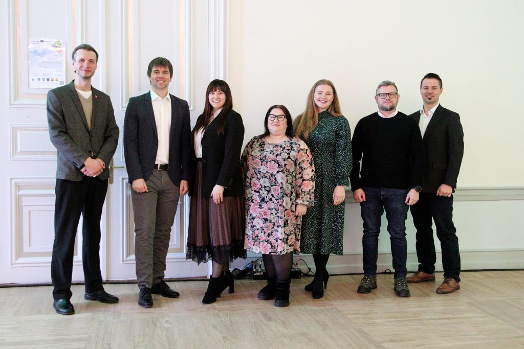 Group photo of seven professionals standing together in a well-lit indoor setting, dressed in formal and semi-formal attire, with a white wall and wooden floor in the background.