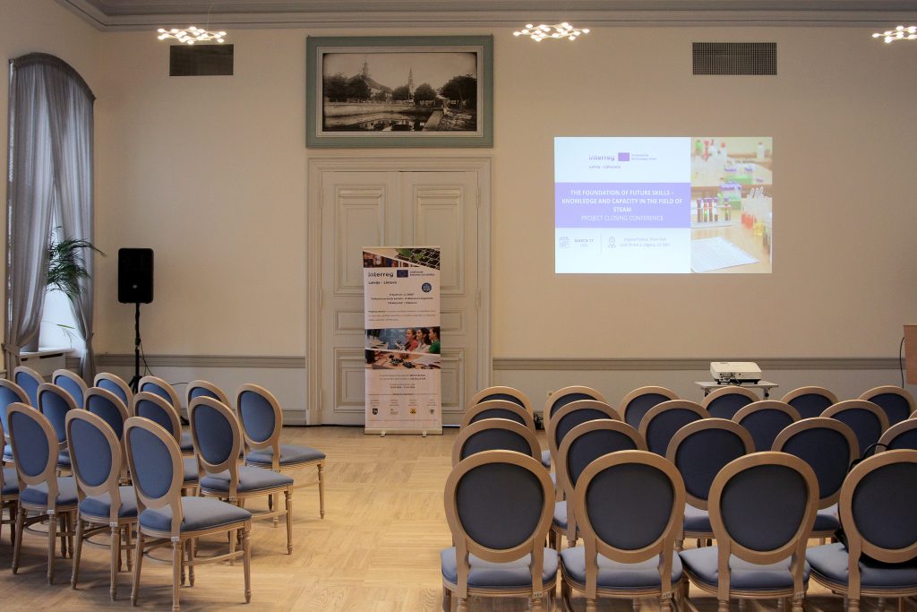 Conference room setup for a project closing event focused on future skills in STEAM. The image shows rows of blue chairs facing a presentation screen displaying event details, along with a banner promoting the Interreg project. The room features elegant decor with natural light and wooden flooring.
