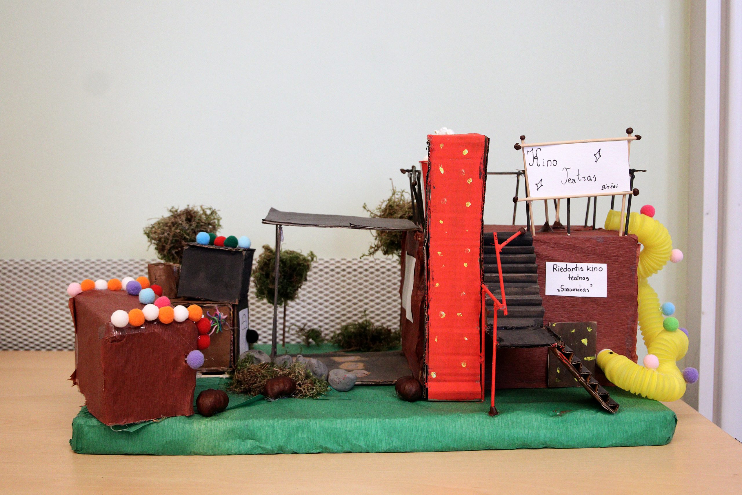 Colorful miniature theater model made from cardboard and decorative materials, featuring a red staircase, signage, and playful embellishments like pom-poms and moss, displayed on a green base.