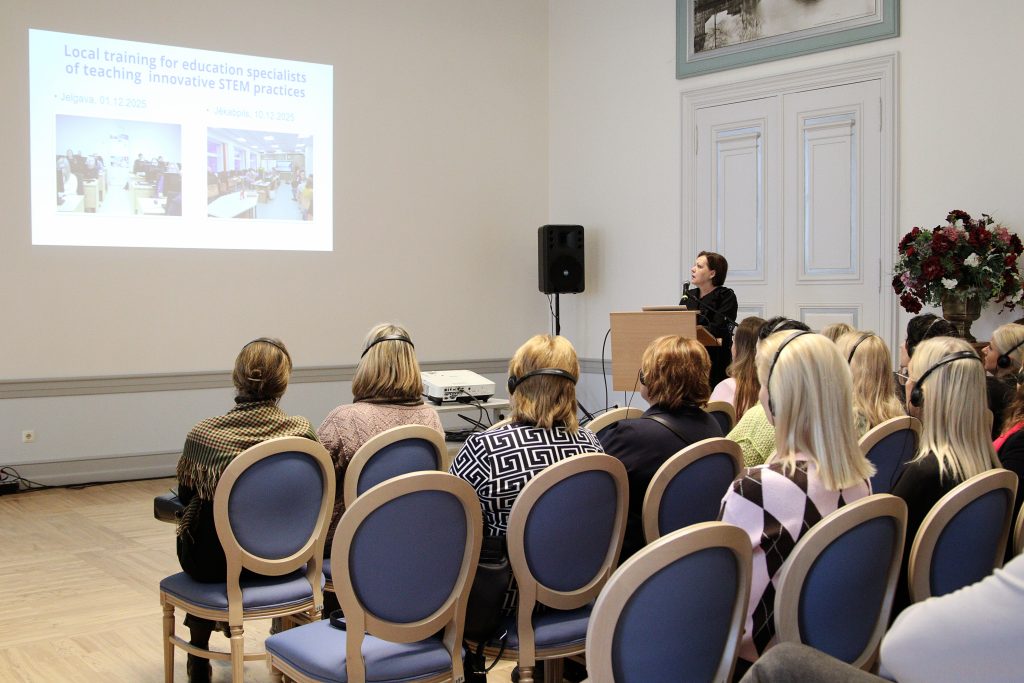 Image of a training session for education specialists on innovative STEM practices, featuring a speaker presenting to an audience. The presentation includes details about local training sessions in Jelgava and Jūkabpils, scheduled for December 2025. Attendees are wearing headsets, indicating a multilingual or specialized training environment.
