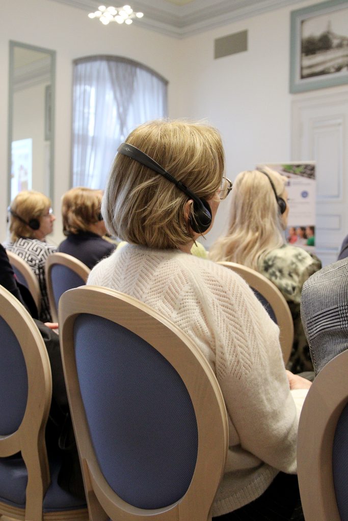 Audience members wearing headphones during a conference or seminar, seated in a well-lit room with modern decor, focusing on the presentation.