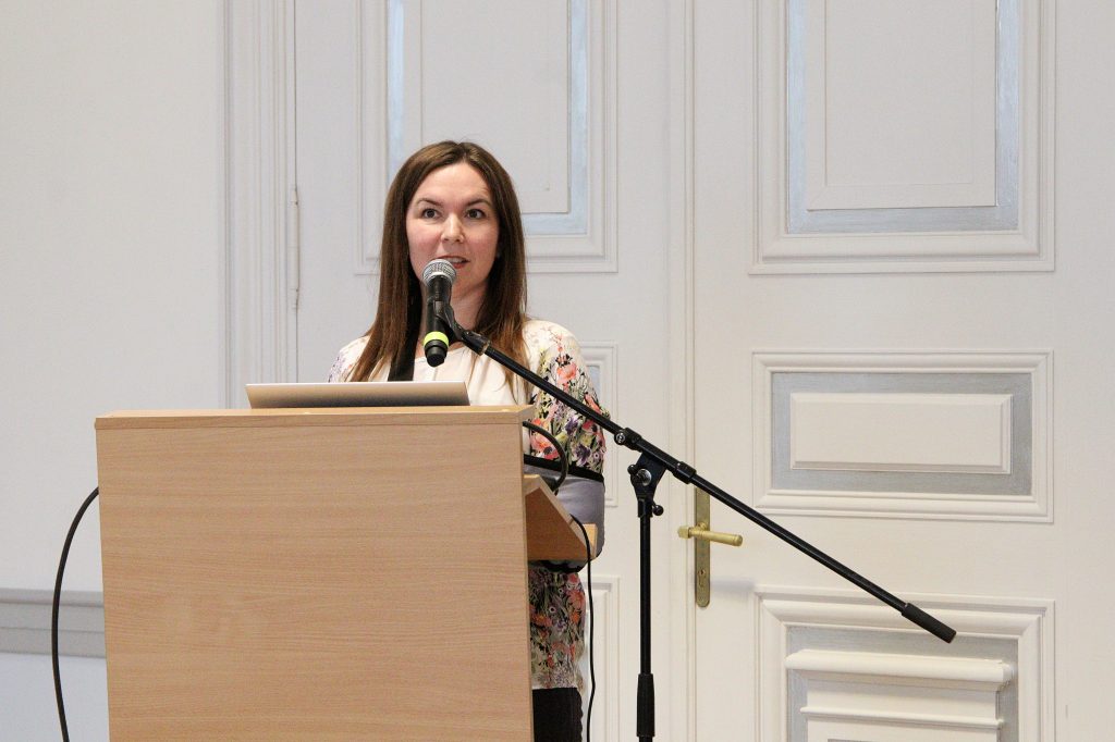 A woman speaking at a podium with a microphone during a presentation, set against a backdrop of elegant white walls.