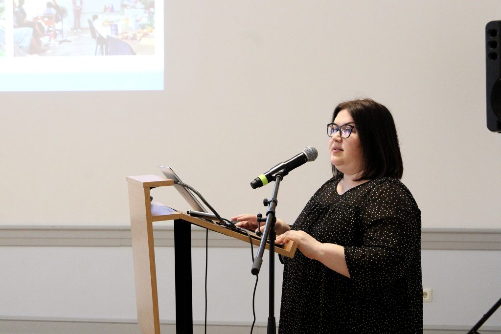 Woman speaking at a podium with a microphone during a presentation, wearing glasses and a black polka dot top, with a laptop and presentation slides visible in the background.