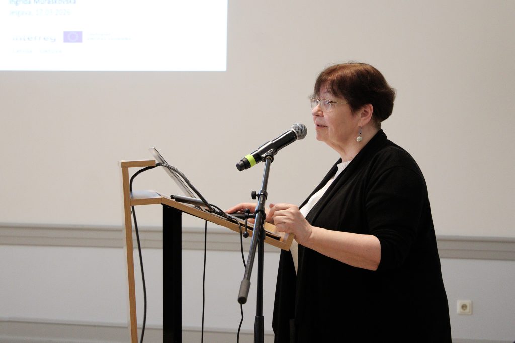 A woman speaking at a podium with a microphone during a presentation, with a projected screen in the background displaying event details in Jelgava, Latvia.