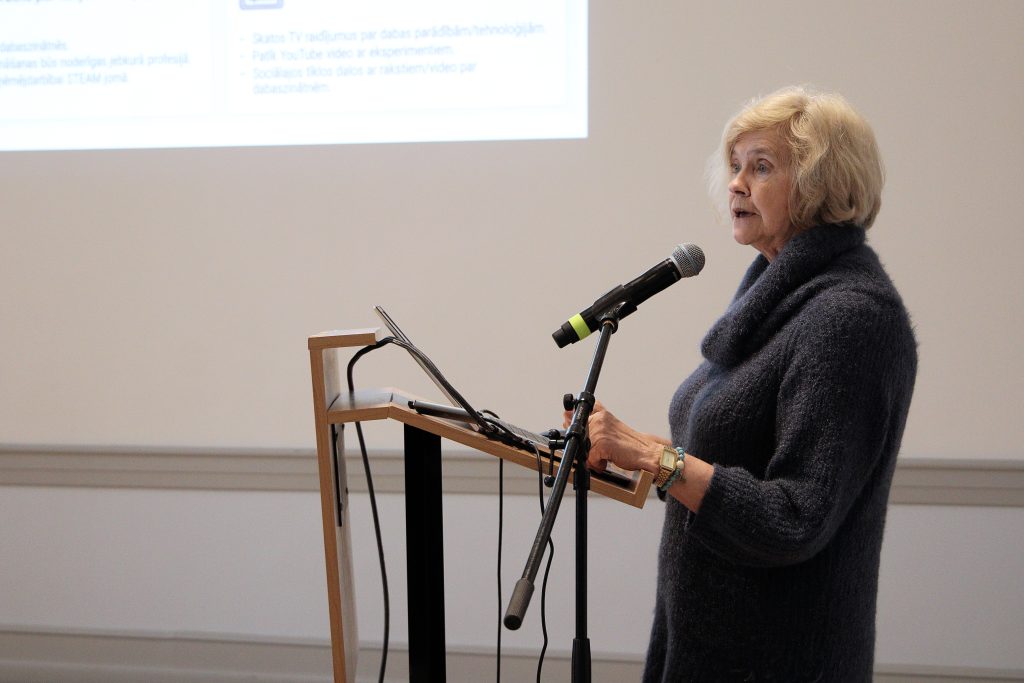 A woman with blonde hair speaks into a microphone at a podium during a presentation, wearing a dark sweater. A presentation screen is visible in the background, displaying text related to data and technology.