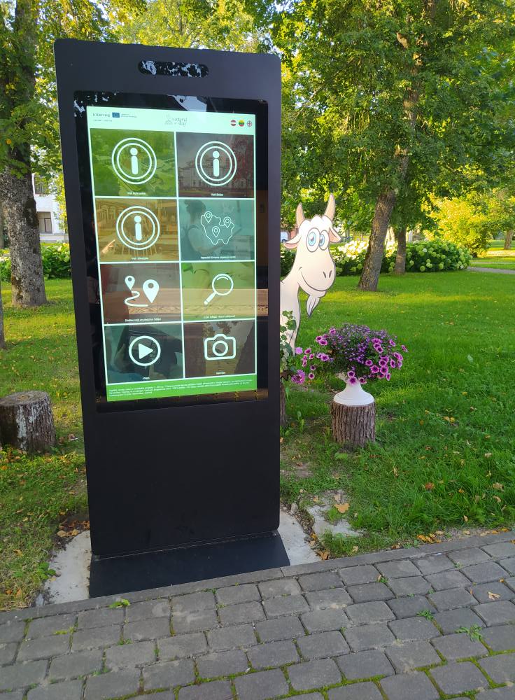 Digital information kiosk in a park displaying various interactive options, with a cartoon goat nearby and colorful flowers in the foreground.