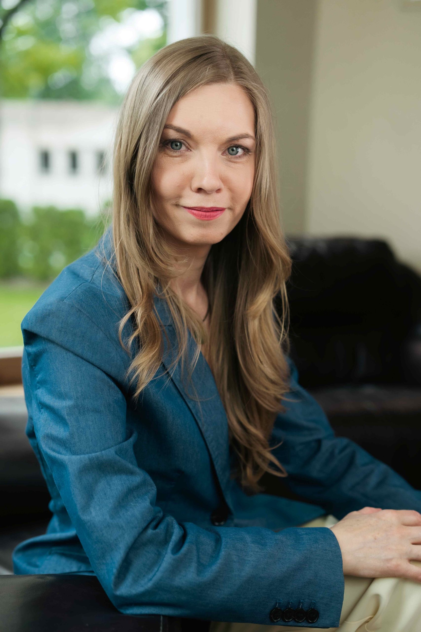 Professional woman with long, wavy hair wearing a blue blazer, seated indoors with a neutral background and natural light.