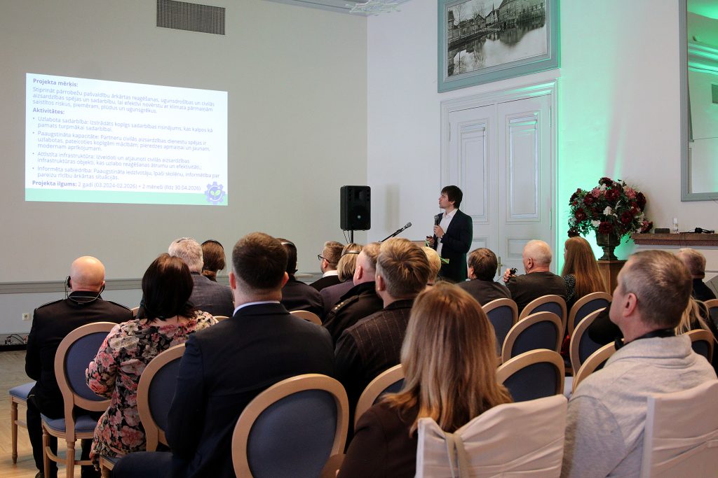 Audience attending a presentation on civil safety and emergency response, featuring a speaker discussing project goals and activities. A PowerPoint slide is visible, outlining objectives and partnership information, in a well-lit conference room setting.