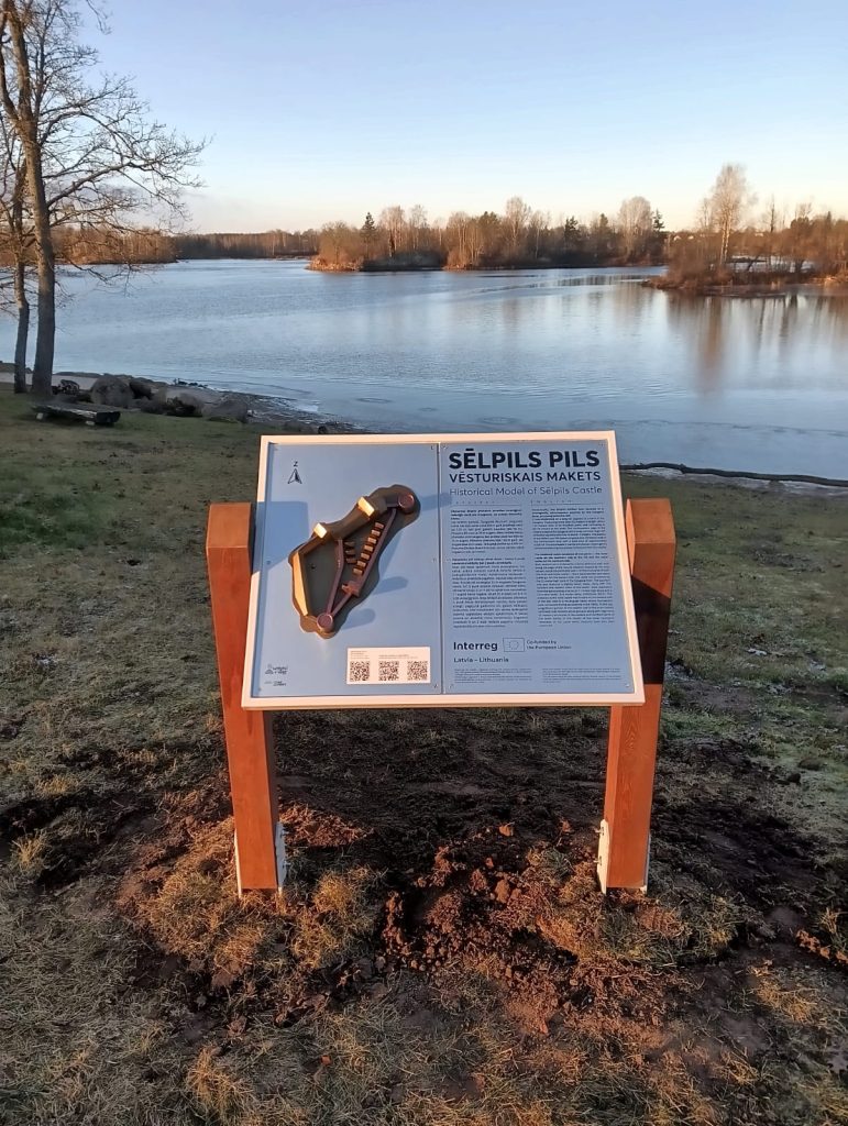 Historical information sign about Sēlpils Castle with a model illustration, set against a serene lakeside view in Latvia, showcasing the natural landscape and cultural heritage.