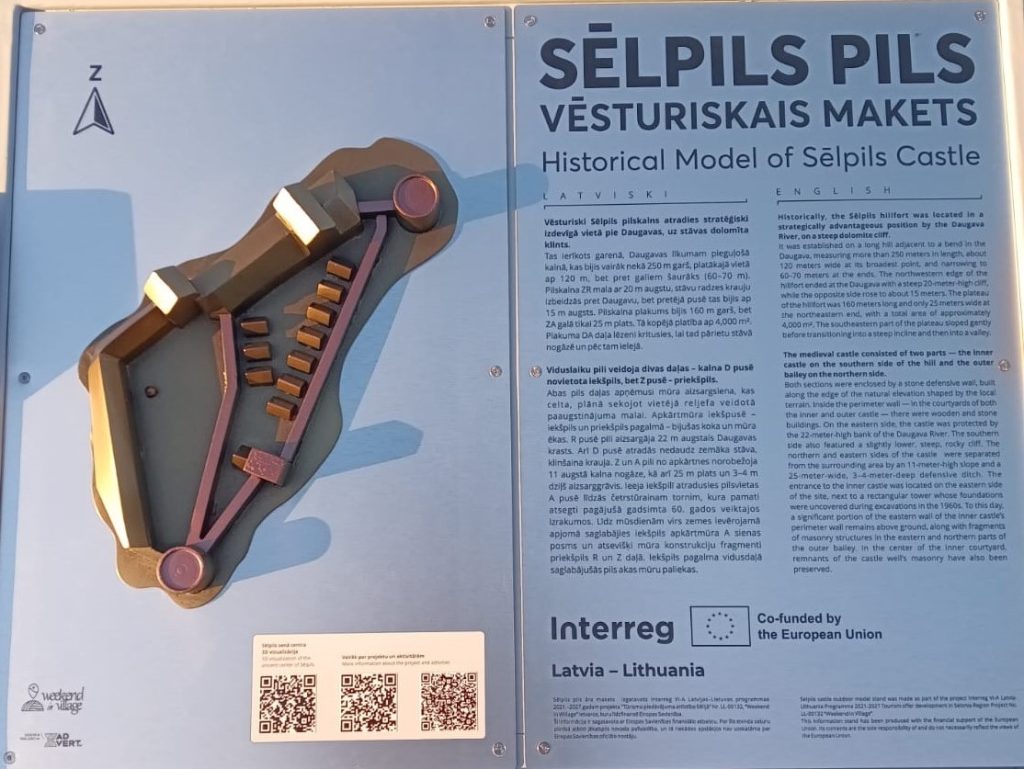 Historical model of Sēlpils Castle displayed on a blue information board, featuring a 3D representation of the castle layout alongside descriptive text in Latvian and English about its strategic location near the Daugava River and historical significance.