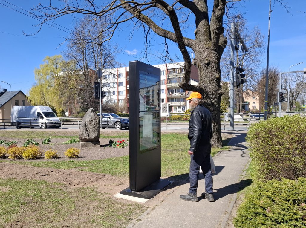 An elderly man in a black leather jacket and orange cap stands near a digital information display in a city park, surrounded by greenery and blooming flowers, with residential buildings and traffic in the background on a sunny day.