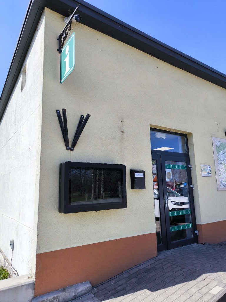 Information center exterior with a green "i" sign, digital display, and a mailbox, set against a clear blue sky. The building features a modern design with a glass door and a map displayed nearby.