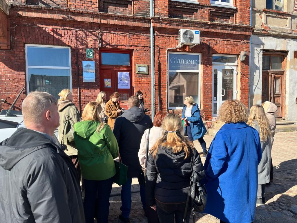 Group of people gathered outside a brick building, engaging in a discussion during a sunny day. The scene features individuals in various outfits, including a blue coat and green jacket, near a storefront with a sign reading "Glamour."