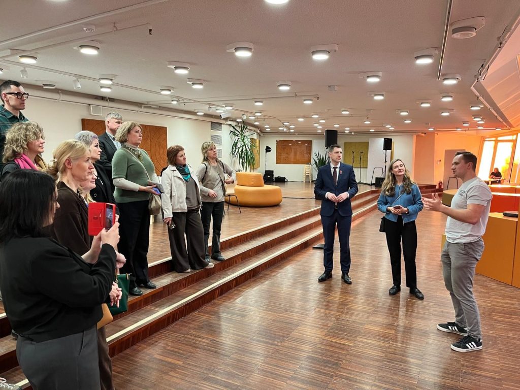 Group of professionals engaged in a discussion during a workshop in a modern event space, featuring a diverse audience listening attentively to a speaker. The setting includes a well-lit room with wooden flooring and contemporary seating arrangements.