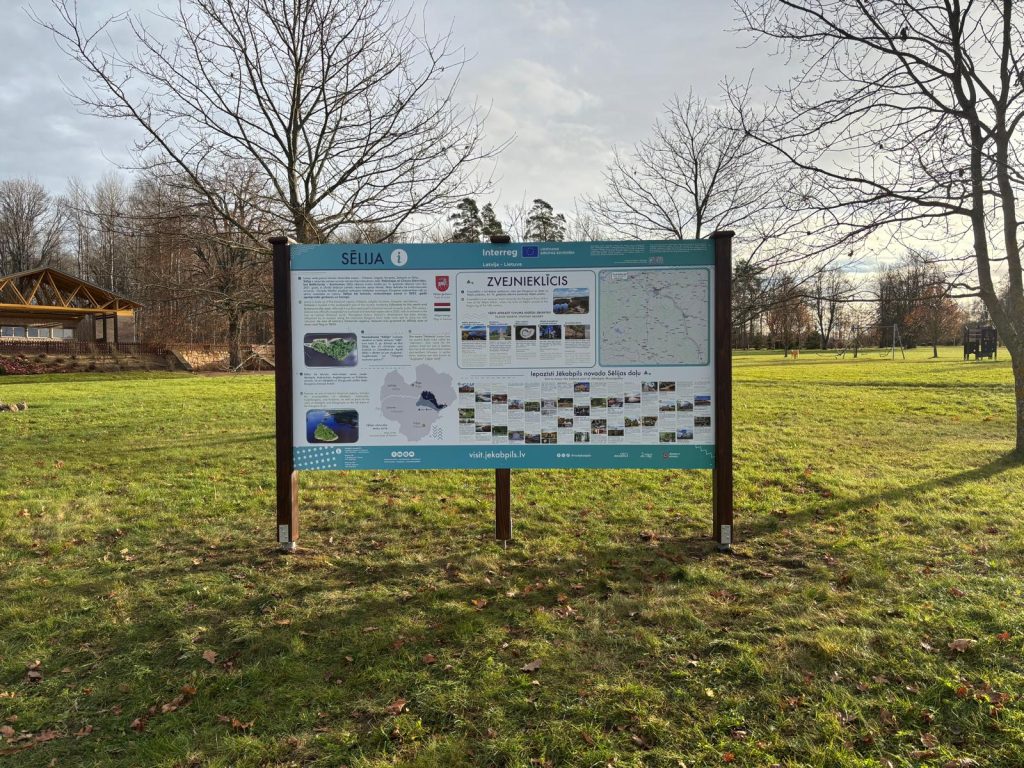 Informative sign about Šļūža and Zvenieki, featuring maps and local attractions, set in a grassy area with trees and a wooden structure in the background. Ideal for visitors seeking information on the region.