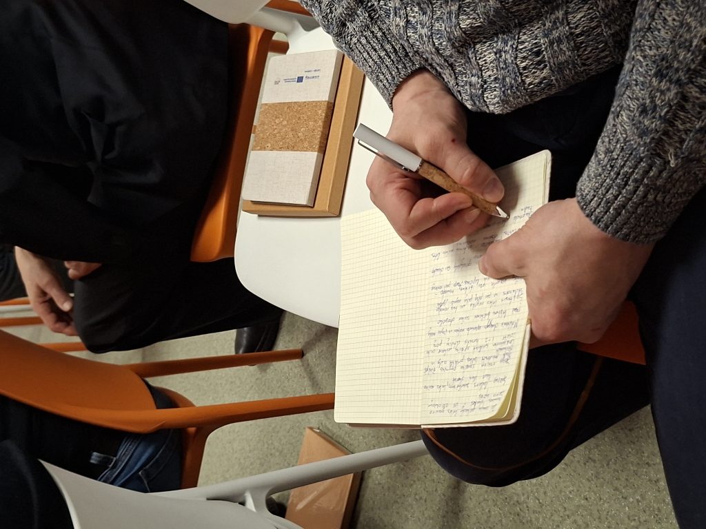 Person taking notes in a notebook during a meeting, with a cork-covered notebook and a pen in hand, seated on an orange chair. Person taking notes in a notebook during a meeting, with a cork-covered notebook and a pen in hand, seated on an orange chair.