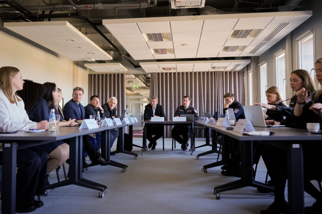 Meeting of professionals in a modern conference room, featuring a diverse group of individuals engaged in discussion around a table, with laptops and nameplates visible. The setting includes large windows and contemporary decor, highlighting a collaborative environment.