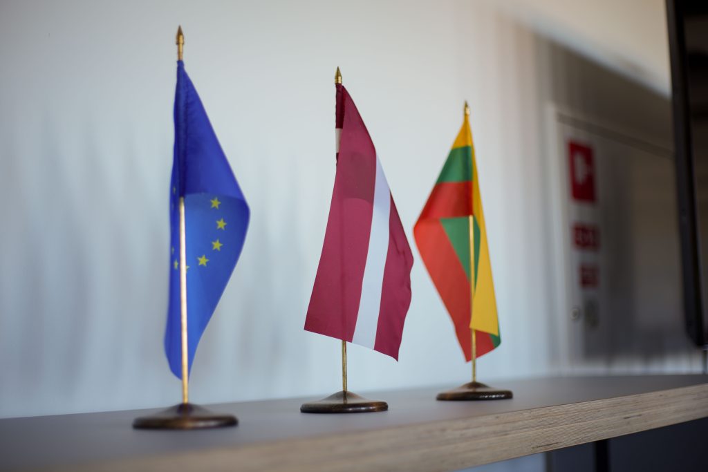 Three small flags are displayed on a wooden shelf, representing the European Union, Latvia, and Lithuania, symbolizing international relations and cooperation.