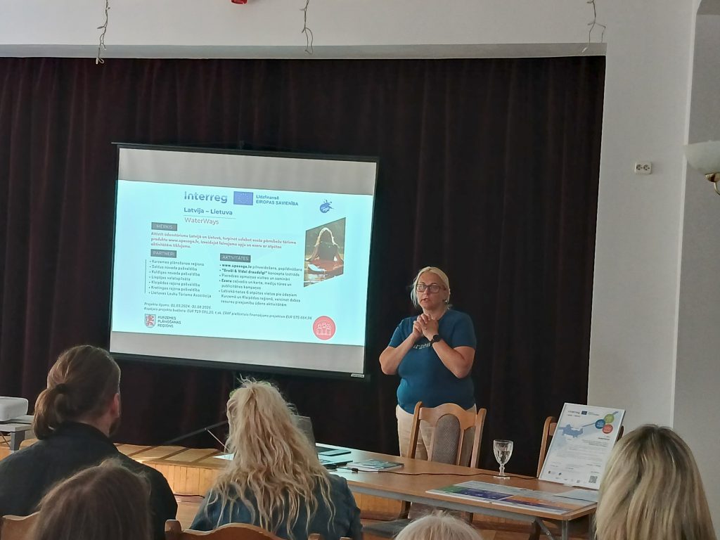 A woman presenting at a conference about the Interreg WaterWays project between Latvia and Lithuania, with a presentation slide visible in the background detailing project goals and activities. Attendees are seated and engaged in the discussion. A woman presenting at a conference about the Interreg WaterWays project between Latvia and Lithuania, with a presentation slide visible in the background detailing project goals and activities. Attendees are seated and engaged in the discussion.
