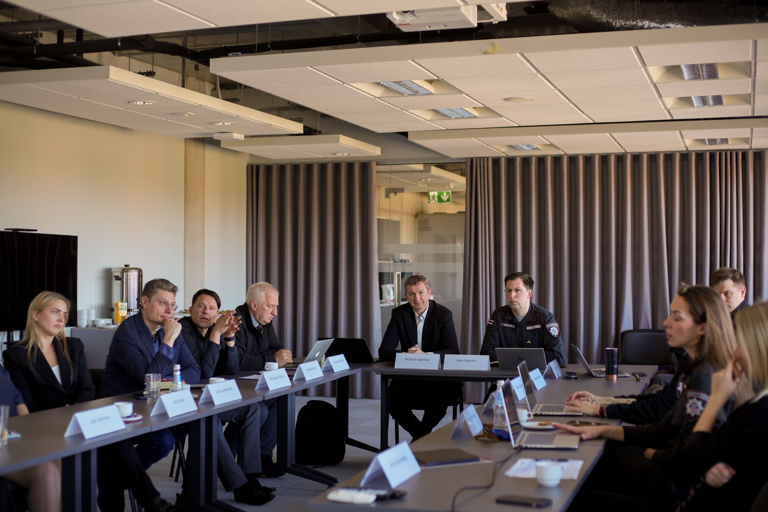 Business meeting in a modern conference room with participants engaged in discussion, featuring laptops and nameplates on a long table, showcasing a collaborative atmosphere among professionals.