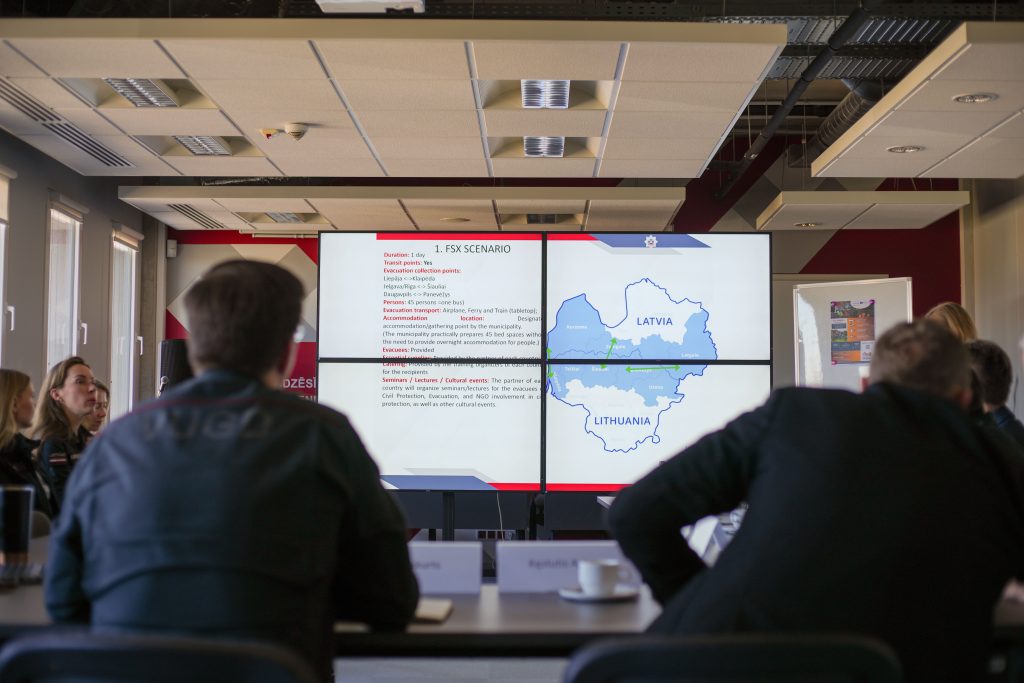 Meeting participants viewing a presentation on emergency evacuation scenarios, featuring a map of Latvia and Lithuania, with details on transit points and accommodation for evacuees displayed on large screens.