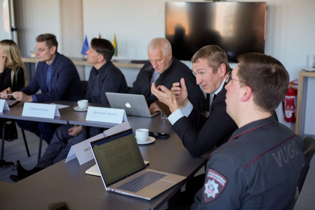 Group of professionals engaged in a discussion around a conference table, featuring laptops and cups of coffee. The setting includes flags in the background, indicating an international or diplomatic context.
