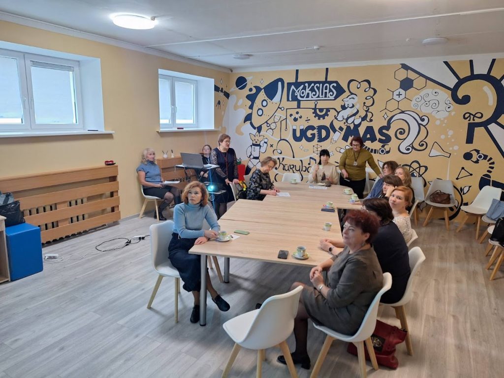 Group of people seated around a large table in a modern meeting room with a colorful mural on the wall, engaged in discussion or a workshop. The setting features bright lighting and contemporary furnishings, creating a collaborative atmosphere.