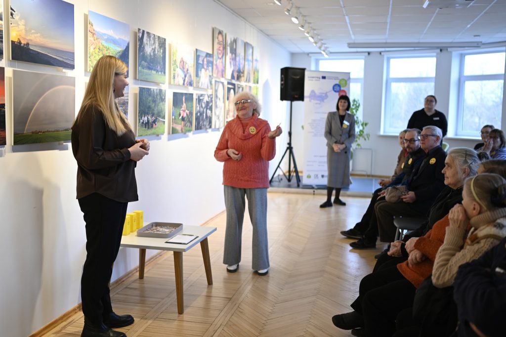 A woman in a black shirt speaks while an elderly woman in an orange sweater gestures during an art exhibition. The gallery features photographs on the walls, and an audience of seniors attentively listens. A table with materials is visible in the foreground. A woman in a black shirt speaks while an elderly woman in an orange sweater gestures during an art exhibition. The gallery features photographs on the walls, and an audience of seniors attentively listens. A table with materials is visible in the foreground.