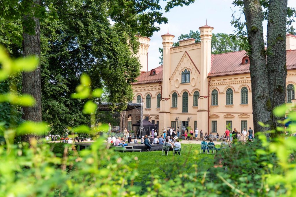 Historic building surrounded by greenery, with people gathered outside for an event. The architecture features ornate details and multiple towers, showcasing a vibrant outdoor atmosphere.