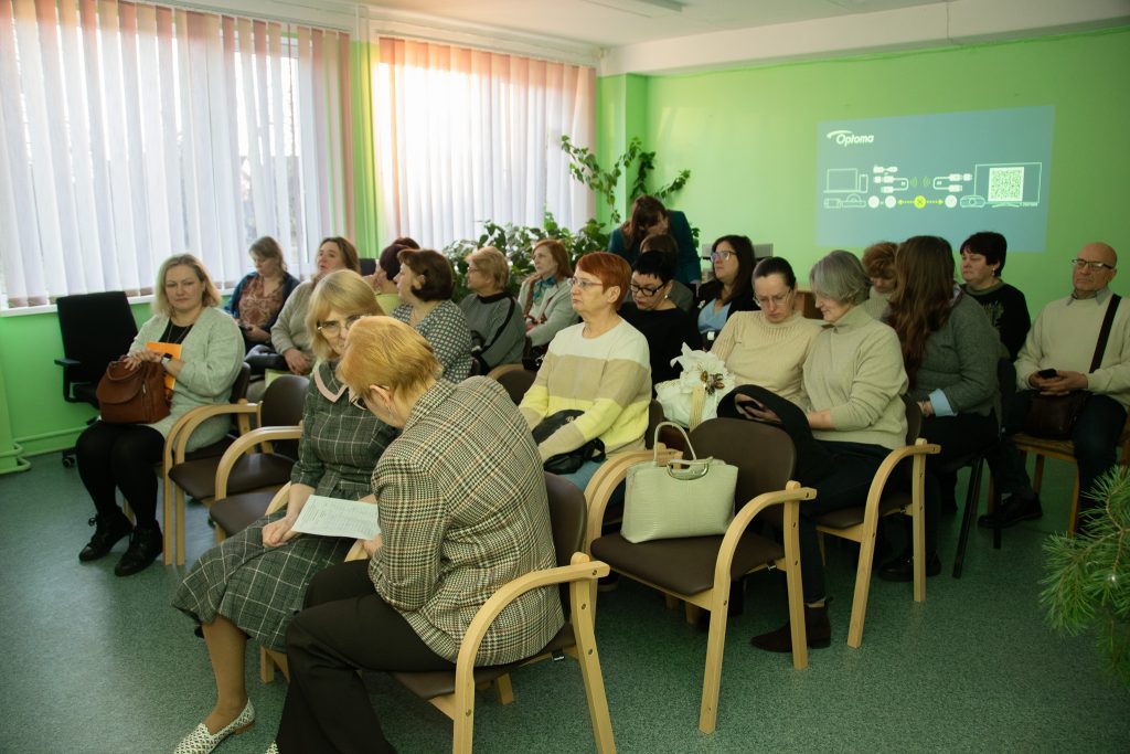 A group of diverse individuals seated in a brightly lit room, engaged in conversation and looking at materials, with green walls and large windows allowing natural light. A presentation is projected on the wall, indicating a professional or community gathering. A group of diverse individuals seated in a brightly lit room, engaged in conversation and looking at materials, with green walls and large windows allowing natural light. A presentation is projected on the wall, indicating a professional or community gathering.
