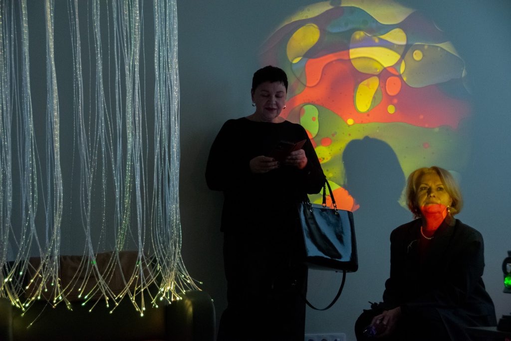 Two women stand in a dimly lit room with colorful abstract projections on the wall. One woman, holding a smartphone, is dressed in black and appears engaged with her device, while the other sits nearby, wearing a dark outfit and looking contemplative. The background features illuminated strands creating a cozy atmosphere. Two women stand in a dimly lit room with colorful abstract projections on the wall. One woman, holding a smartphone, is dressed in black and appears engaged with her device, while the other sits nearby, wearing a dark outfit and looking contemplative. The background features illuminated strands creating a cozy atmosphere.