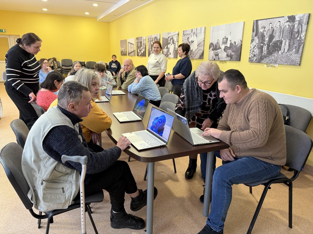 Group of people engaged in a technology workshop, learning on laptops in a community center with yellow walls and historical photographs in the background. Group of people engaged in a technology workshop, learning on laptops in a community center with yellow walls and historical photographs in the background.