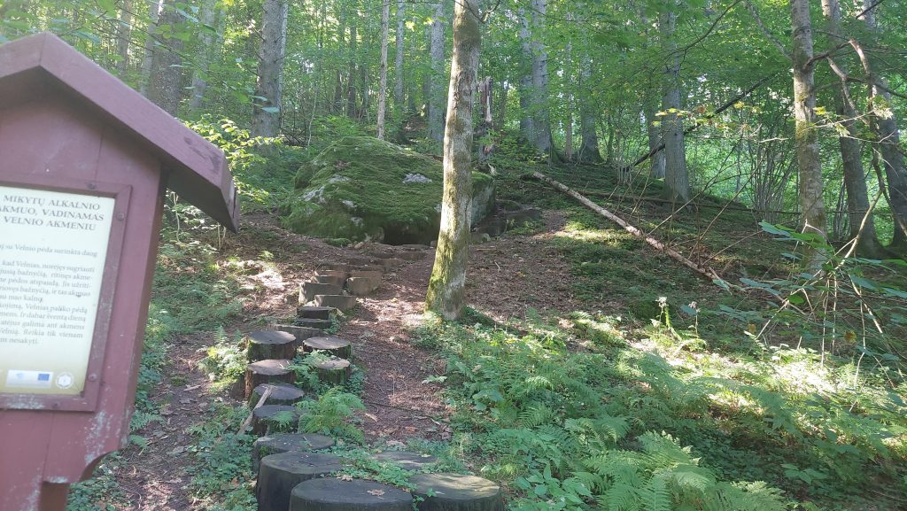 Pathway leading through a lush green forest with moss-covered rocks and wooden steps, featuring an informational sign about the legend of the Devil's footprint in Mikytai Alkalnio. Pathway leading through a lush green forest with moss-covered rocks and wooden steps, featuring an informational sign about the legend of the Devil's footprint in Mikytai Alkalnio.