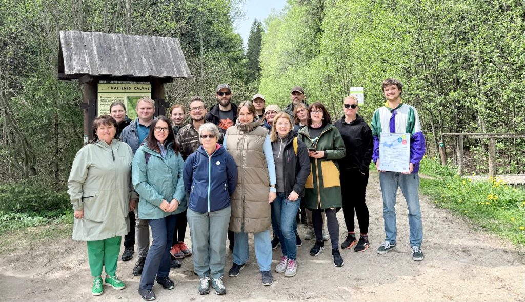 Group photo of participants at Kaltenes Kalvas nature site, showcasing diverse individuals in outdoor attire, gathered near an information sign in a lush green forest setting.