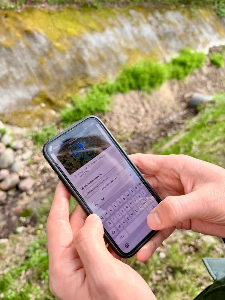 Person using a smartphone to input location details into a mapping application, with a natural background featuring grass and a stone wall.