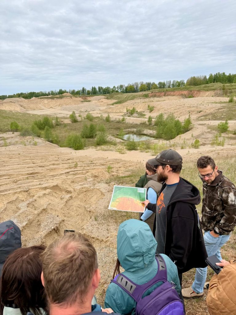 Group of people observing a sand extraction site while discussing a topographic map, surrounded by a natural landscape with sparse vegetation and cloudy skies.