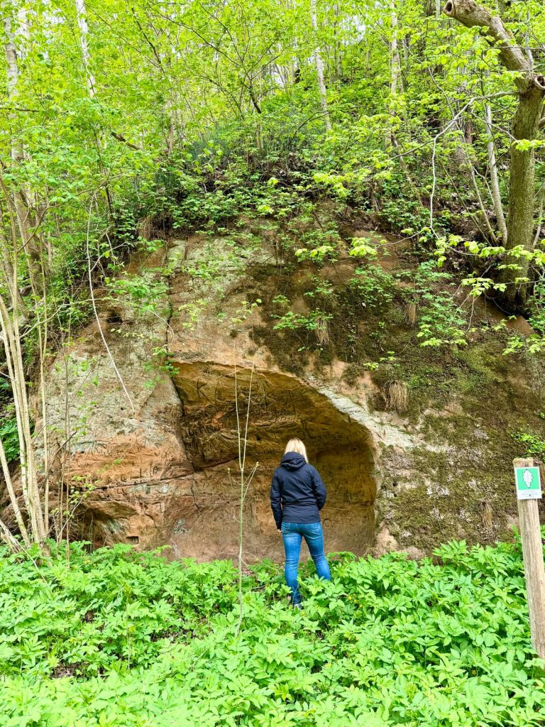 Person standing in front of a natural rock formation surrounded by lush green foliage and trees, exploring the scenic landscape.