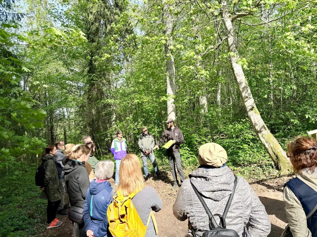 Group of people gathered in a lush green forest for an educational nature walk, listening to a guide presenting information about the local ecology and conservation efforts.