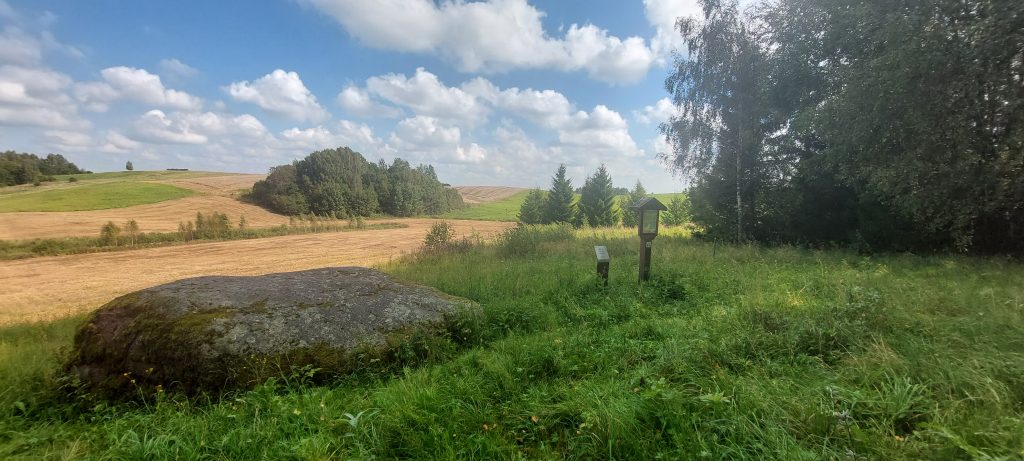 Scenic rural landscape featuring a large rock, grassy area, and wooden signage amidst rolling hills and a blue sky with clouds. Ideal for nature walks and outdoor exploration. Scenic rural landscape featuring a large rock, grassy area, and wooden signage amidst rolling hills and a blue sky with clouds. Ideal for nature walks and outdoor exploration.