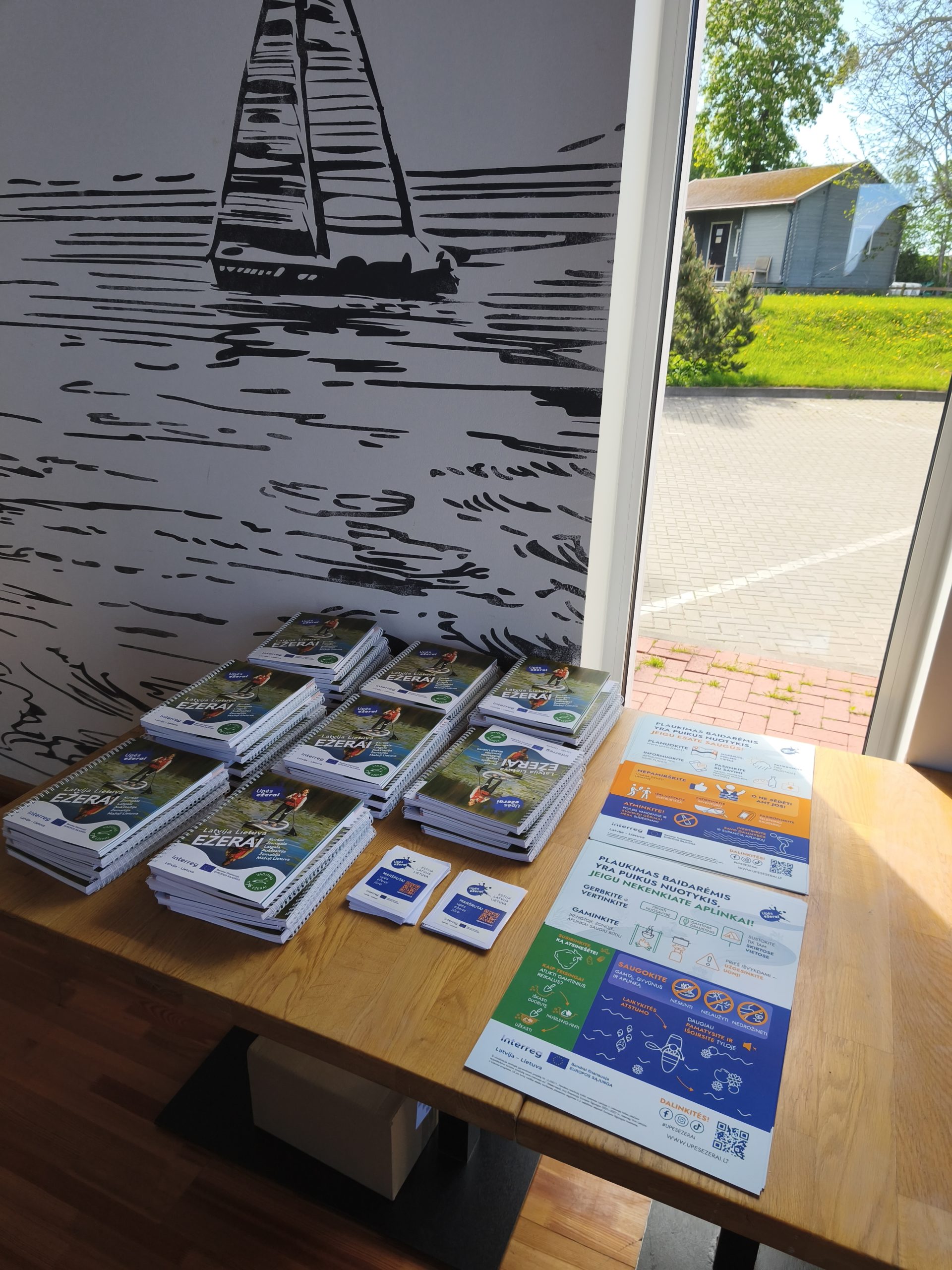 Display of informational brochures and materials on a wooden table, featuring spiral-bound booklets and flyers related to environmental initiatives. The background includes a stylized mural of a sailboat on water, with a view of greenery and a building outside the window.