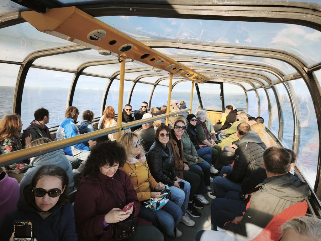 Interior view of a sightseeing boat filled with passengers enjoying a scenic ride over water, featuring large windows and a bright atmosphere.