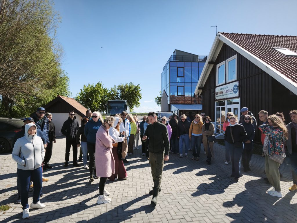 Group of people gathered outside a building with a sign for "Strykis Žuvies Turgus," enjoying a sunny day. The scene features a diverse crowd, including individuals in casual attire, standing on a paved area with trees and vehicles in the background.